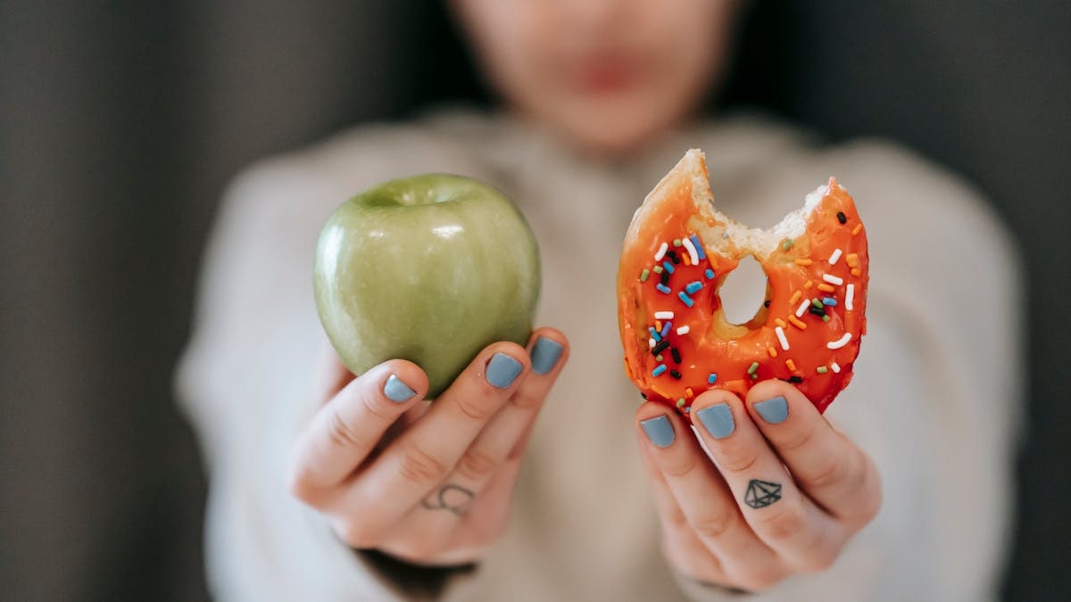 Crop anonymous female demonstrating delicious sweet donut and fresh green apple while making decision